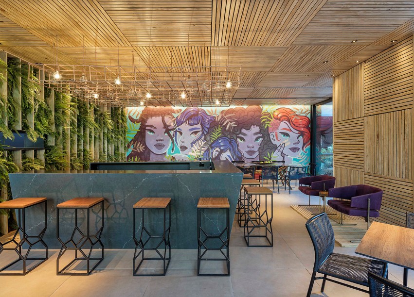 Pizza Bar – Studio Gontijo Arquitetura e Interiores. Criado por mulheres para uma chef, Narciza Leão, chama a atenção inicialmente pelo grafite da artista brasiliense Camila Siren. Outro elemento importante é o deck de plástico reciclado, aplicado em todo o teto e em uma das paredes. Do lado oposto, fica o jardim vertical com a espécie Barba de Velho. No alto, a luminária de cobre não passa despercebida, leve e geométrica.