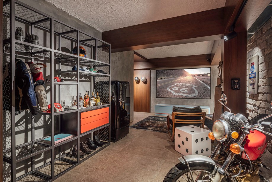 Garagem do Motociclista - Marcelo Oliveira. O espaço de 25 m² traz a irreverência dos anos 1970, a meio caminho entre uma sala de estar e um porão. As poltronas Pitu, do designer Aristeu Pires, trazem conforto para ver filmes e jogar vídeo-game. A tecnologia de automação contrasta os materiais brutos, como pedras naturais, cimento aparente, MDF madeirado e tijolos.