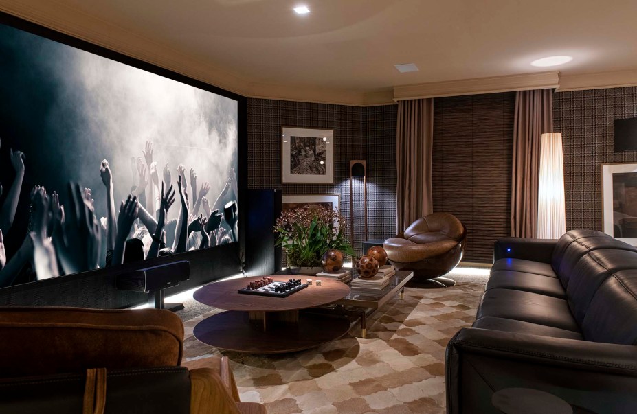 Home Theater – Marlon Gama. O ambiente assinado pelo arquiteto Marlon Gama é dividido em três espaços distintos — espaço para TV com sofá e poltronas; espaço de jogos; e ambiente de bar para total interação da família. Marlon estudou a reverberação do ambiente a fim de que o conforto acústico fosse maximizado. Para tanto, se valeu de tecidos nos revestimentos das paredes, do uso do couro no sofá e nas poltronas, da madeira texturizada e de um tapete que recobre toda a extensão do ambiente. A coleção de obras de arte contemporânea exibe nomes como Artur Lescher, Vik Muniz, Fabi Aleixo, Carybé e Floriano Teixeira. Destaque: Escultura de Ascânio do acervo do arquiteto.