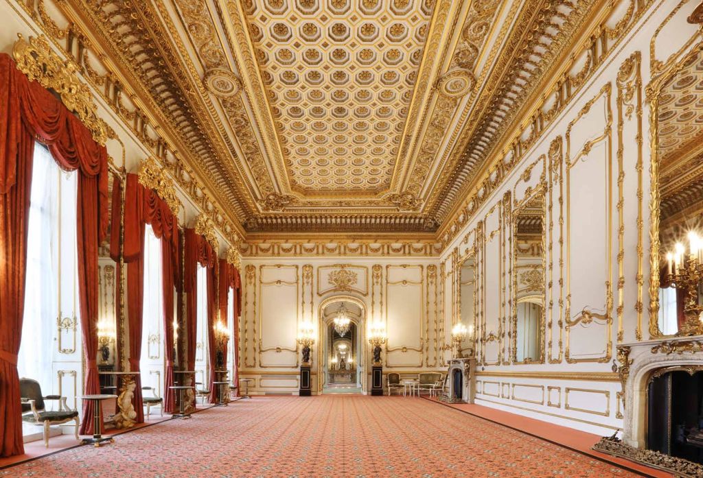 Interior de sala de palácio com tapete vermelho, teto e paredes ornamentadas com detalhes dourados. Janelas altas com cortinas vermelhas