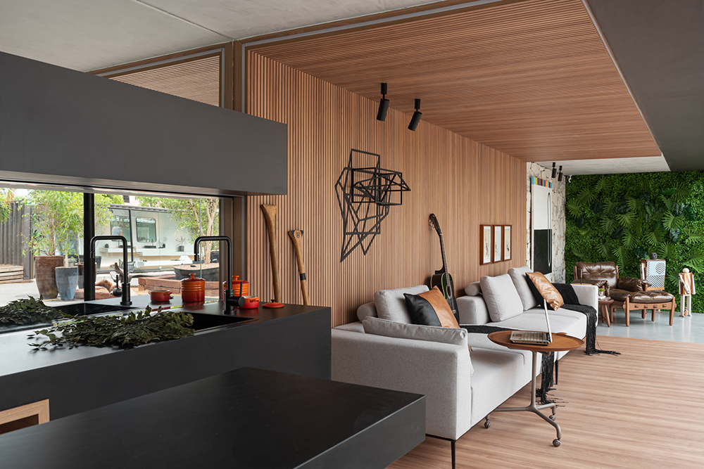 LETÍCIA FINAMORE _ CORAÇÃO DA CASA _ CSF_3120 Living com revestimento em madeira. Sofás cinzas e parede verde ao fundo. Cozinha com bancada preta integrada.