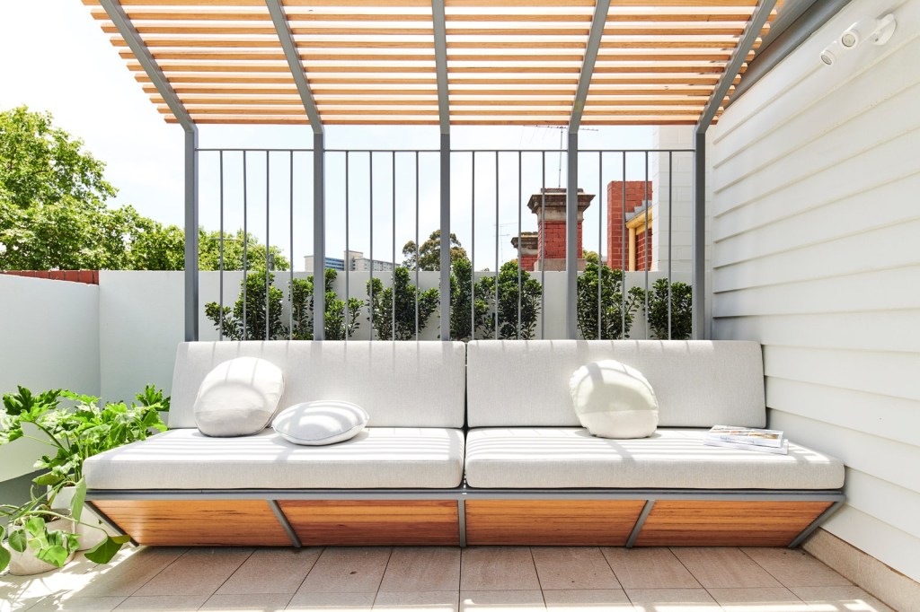 Banco ao ar livre, com suporte de madeira e estofado em branco, coberto por um gazebo de madeira. Ao fundo, plantar verdes.