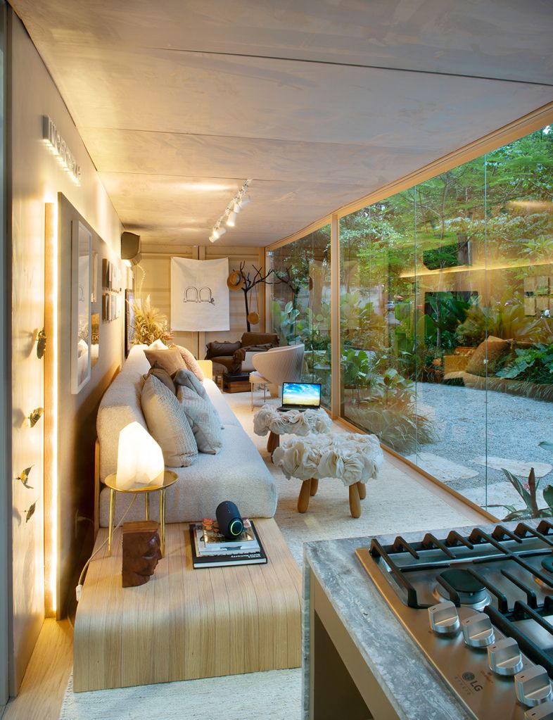 Living com mesa lateral, painel iluminado, sofá e banquinhos de apoio; à frente a parede de vidro do contêiner 