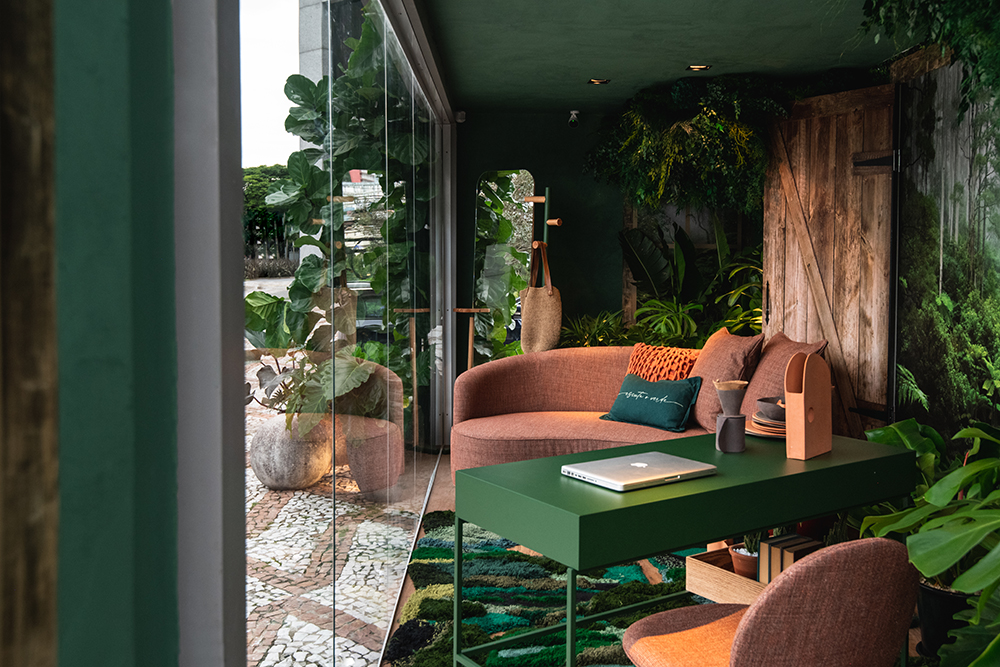 Imagem do home office com sofá laranja e mesa verde envolto por plantas