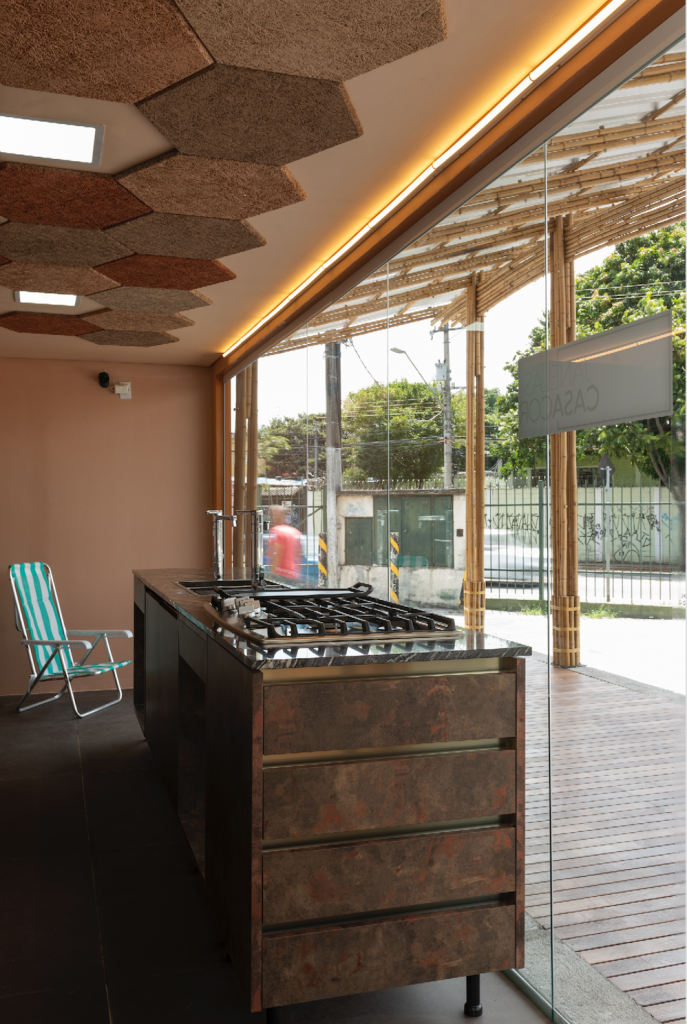 faud-acolhe-cria-cozinha-comunitaria-em-brasilandia-no-janelas-casacor-sp – 10 Parte interna da cozinha comunitária, com a bancada em tom de ferrugem, com um cooktop e torneira. Ao fundo, uma cadeira de praia com listras azuis e brancas. No teto, peças hexagonais e na parede marrom avermelhado. Do lado de fora, atrás do vidro do conteiner, um deck de piso de madeira, e cobertura feira com estrutura de bambu.