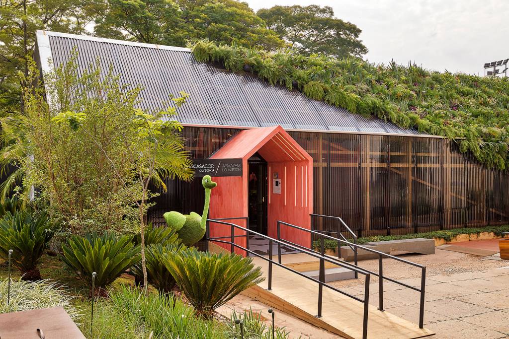 Portal com topo triangular em vermelho. O telhado verde e coberto até a metade em uma linha diagonal. À esquerda da entrada, um avestruz de planta.