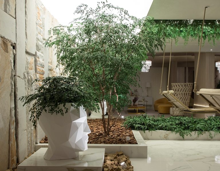 urban-jungle-CasaCorSC-paisagismo Plantas na área externa de um empreendimento presentes em uma pequena árvore e um vaso em formato de cabeça humana geométrico que abriga plantas também