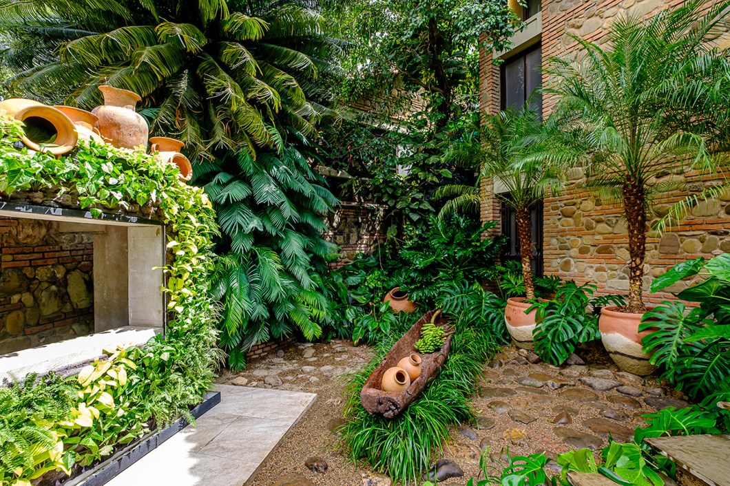 Jardín de Piedra - Natalia Murillo. Para valorizar a arquitetura original da casa, a paisagista utilizou diversos materiais naturais em seu jardim - como argila, pedra e palha - que também ressaltam a natureza existente. “No meio da nossa vegetação composta por guaimbês nativos, filodendros e costelas-de-adão, abrimos espaço para uma imponente canoa chiquitana carregada de frutos de nossa terra”, detalha a profissional. O verde também envolve a bilheteria, enquadrada por jardins verticais. Jardín de Piedra - Natalia Murillo. Para valorizar a arquitetura original da casa, a paisagista utilizou diversos materiais naturais em seu jardim - como argila, pedra e palha - que também ressaltam a natureza existente. “No meio da nossa vegetação composta por guaimbês nativos, filodendros e costelas-de-adão, abrimos espaço para uma imponente canoa chiquitana carregada de frutos de nossa terra”, detalha a profissional. O verde também envolve a bilheteria, enquadrada por jardins verticais.