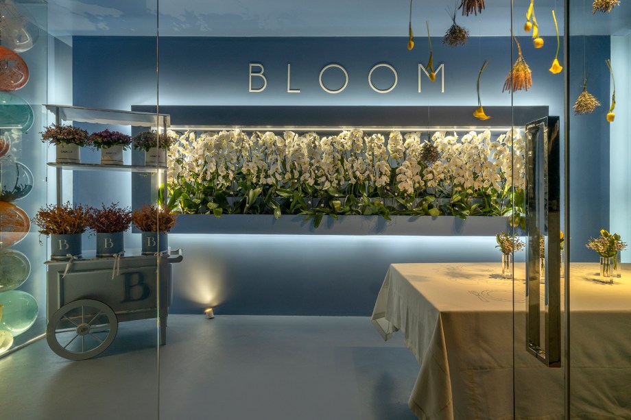 Roberto Guedes - Bloom. Um dos ambientes mais instagramáveis da mostra, Roberto Guedes projetou uma parede de balões e flores excêntricas. Iluminação de led dá o charme ao ambiente minimalista. A inspiração do arquiteto foi em lojas italianas ligadas à área de moda e flores, após uma longa pesquisa na forma como estabelecimentos desses nichos apresentam suas criações. Com 18 m² em construção modular, o ambiente utiliza a cor azul tibet. Usando a mesma cor em todo o ambiente com o intuito de levar o visitante a valorizar apenas um momento, o espaço traz ornamentação com pratos exclusivos, vasos e flores.