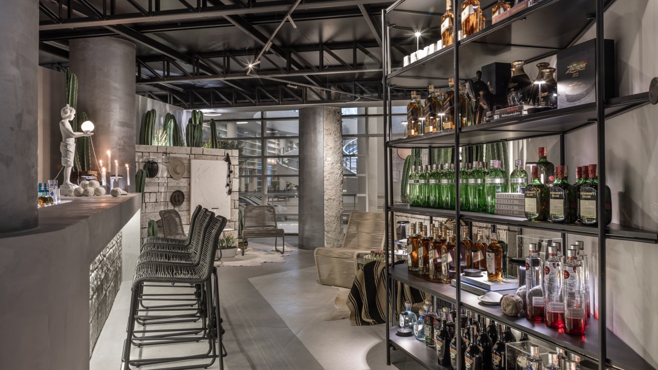 Eduardo Baldelomar - UYUNI Lounge The - Bar. Projeto da CASACOR São Paulo 2023. Na foto, bar com balcão de pedra iluminado, estante com bebidas e obras de arte.