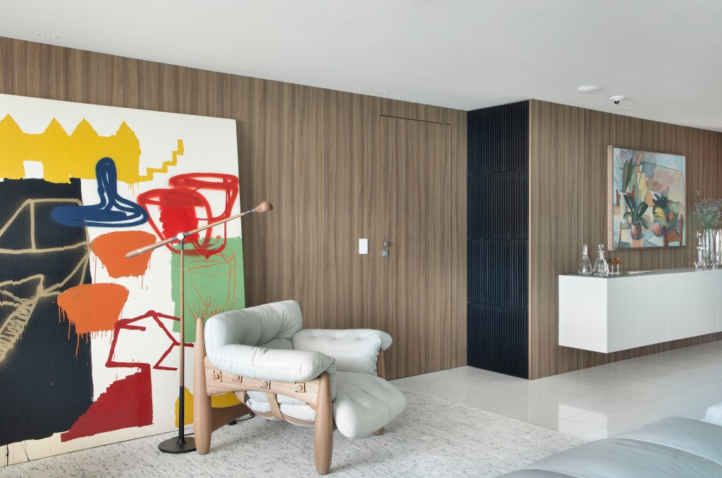 decor-ape-200-m2-valoriza-vista-mar-obras-de-arte-ana-moura-ana-higino-credito-denilson-machado (15) Décor de apê de 200 m² valoriza a vista do mar e obras de arte da moradora. Projeto de Ana Moura Ana Higino Arquitetura. Na foto, sala com parede de madeira, porta camuflada e obras de arte.