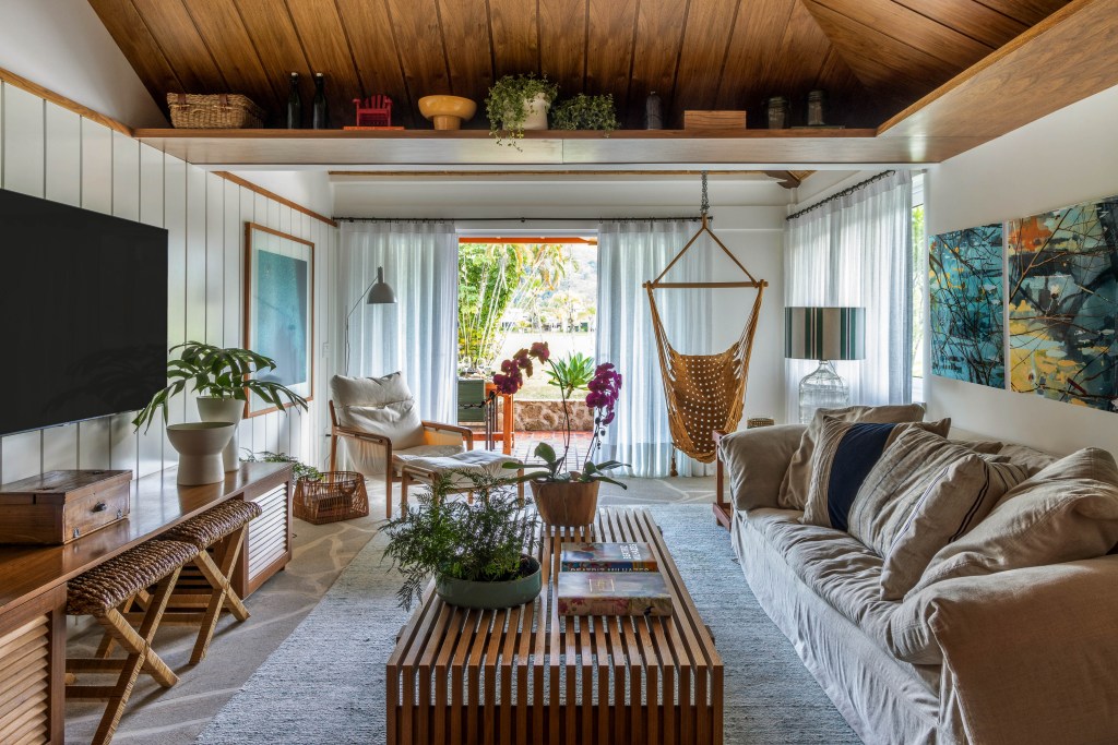 Casa de condomínio de 110 m² tem vista para o canal e décor "praia chic". Projeto de Paola Ribeiro. Na foto, sala de estar com móveis de madeira, poltrona suspensa e vista para a varanda.