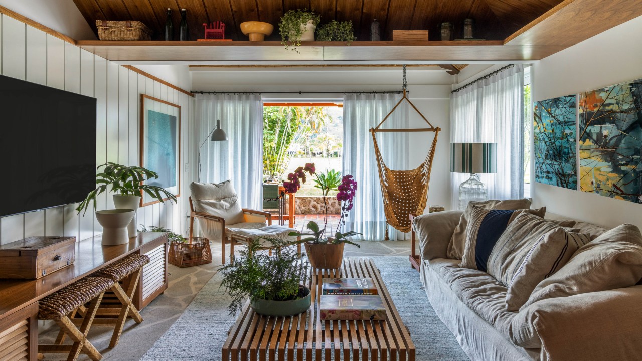 Casa de condomínio de 110 m² tem vista para o canal e décor "praia chic". Projeto de Paola Ribeiro. Na foto, sala de estar com móveis de madeira, poltrona suspensa e vista para a varanda.
