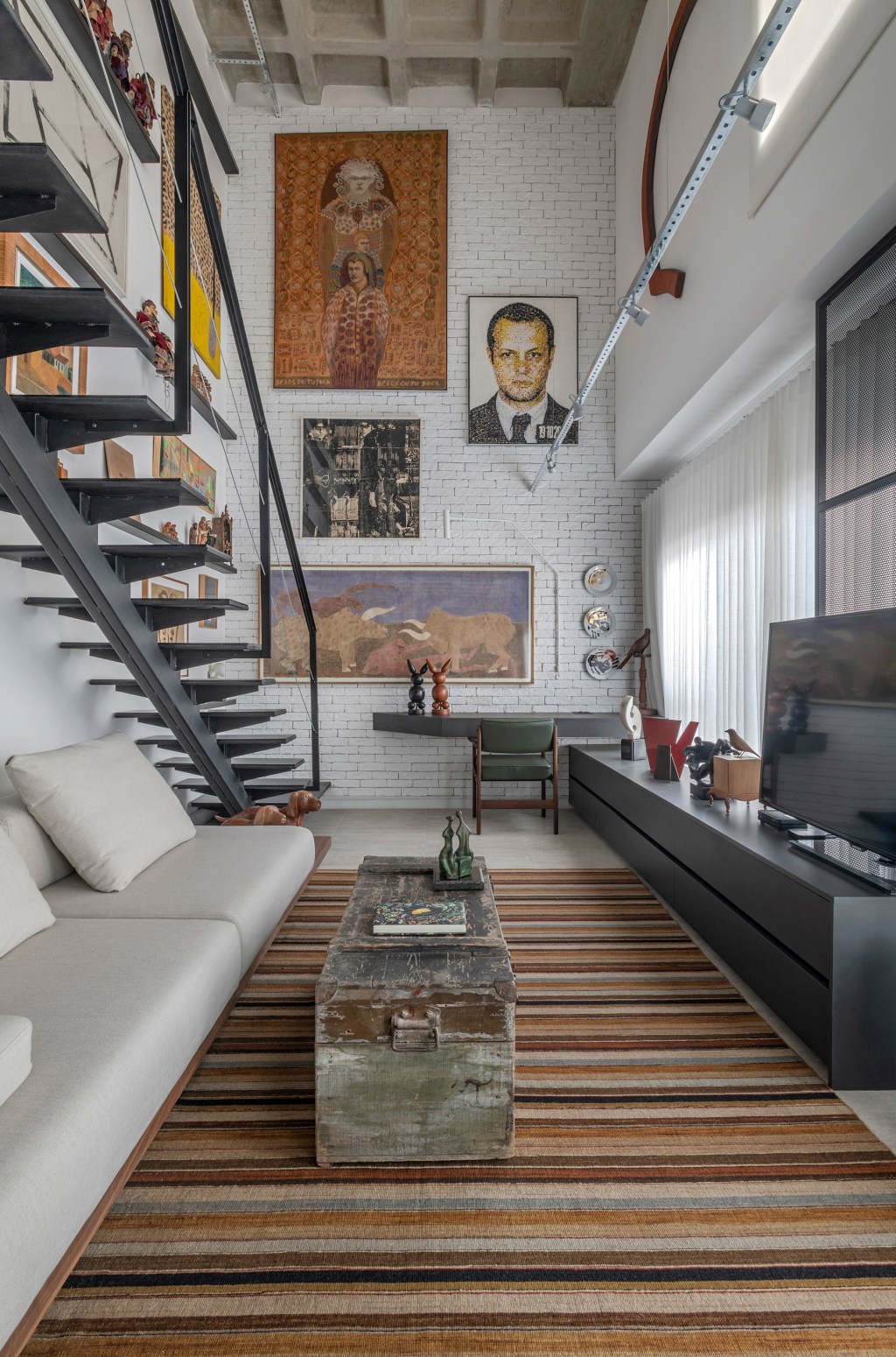 Loft de 64 m² ganha décor industrial e coleção de arte dos moradores. Projeto de Junior Piacesi. Na foto, escada com gallery wall na parede. Sala de estar com tapete listrado.