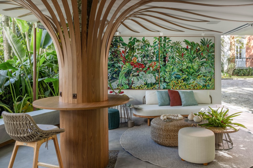 Ana Cano e Vivian Reimers - Lounge das boas-vindas. O Lounge das Boas-Vindas, junto à bilheteria, foi criado pelas arquitetas Ana Cano e Vivian Reimers como um lugar de descanso e encontro com os amigos. O ambiente aposta na biofilia com um décor repleto de elementos naturais como o bambu, que aparece nos revestimentos; o barro reciclado, que cobre os nichos da estante existente; pedras e tramas. Uma grande árvore se destaca ao centro do espaço que segue a paleta de cores do jardim com verdes e terrosos e tem mobiliário de formas orgânicas. Ana Cano e Vivian Reimers - Lounge das boas-vindas. O Lounge das Boas-Vindas, junto à bilheteria, foi criado pelas arquitetas Ana Cano e Vivian Reimers como um lugar de descanso e encontro com os amigos. O ambiente aposta na biofilia com um décor repleto de elementos naturais como o bambu, que aparece nos revestimentos; o barro reciclado, que cobre os nichos da estante existente; pedras e tramas. Uma grande árvore se destaca ao centro do espaço que segue a paleta de cores do jardim com verdes e terrosos e tem mobiliário de formas orgânicas.