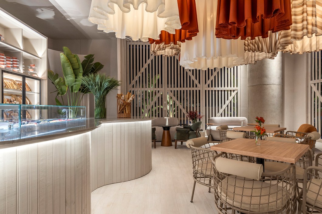 Karla Amaral - Boulangerie. O espaço de 110 m² foi idealizado oor Karla Amaral para ser um refúgio para os dias agitados. A cor branca foi colocada como base do projeto para criar um ambiente luminoso. Materiais de diferentes texturas associados ao verde da vegetação imprimem aconchego e intimidade. Os brises, que são referências do modernismo candango, foram ressignificados através de enormes portas pivotantes em madeira estilo celeiro, o que eleva o olhar para o ponto focal no salão de mesas: as luminárias desenhadas especialmente para o café. Karla Amaral - Boulangerie. O espaço de 110 m² foi idealizado oor Karla Amaral para ser um refúgio para os dias agitados. A cor branca foi colocada como base do projeto para criar um ambiente luminoso. Materiais de diferentes texturas associados ao verde da vegetação imprimem aconchego e intimidade. Os brises, que são referências do modernismo candango, foram ressignificados através de enormes portas pivotantes em madeira estilo celeiro, o que eleva o olhar para o ponto focal no salão de mesas: as luminárias desenhadas especialmente para o café.