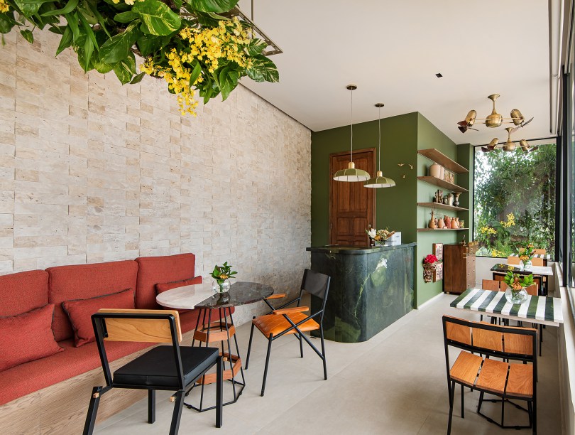 Gabriella Machado Arquitetura - Cafeteria das Lembranças. A profissional buscou ressaltar a conexão com a natureza por meio do uso de materiais em seu estado natural, como a pedra rústica da parede e a madeira. As cores desempenham um papel essencial na ambientação: o vermelho e o verde foram inspirados nas tonalidades do fruto e do ramo do café e aparecem no sofá, na parede e na arte da fachada. As mesas são feitas a partir de sobras de mármores e de gradis antigos e a porta de madeira foi resgatada de um palacete histórico.