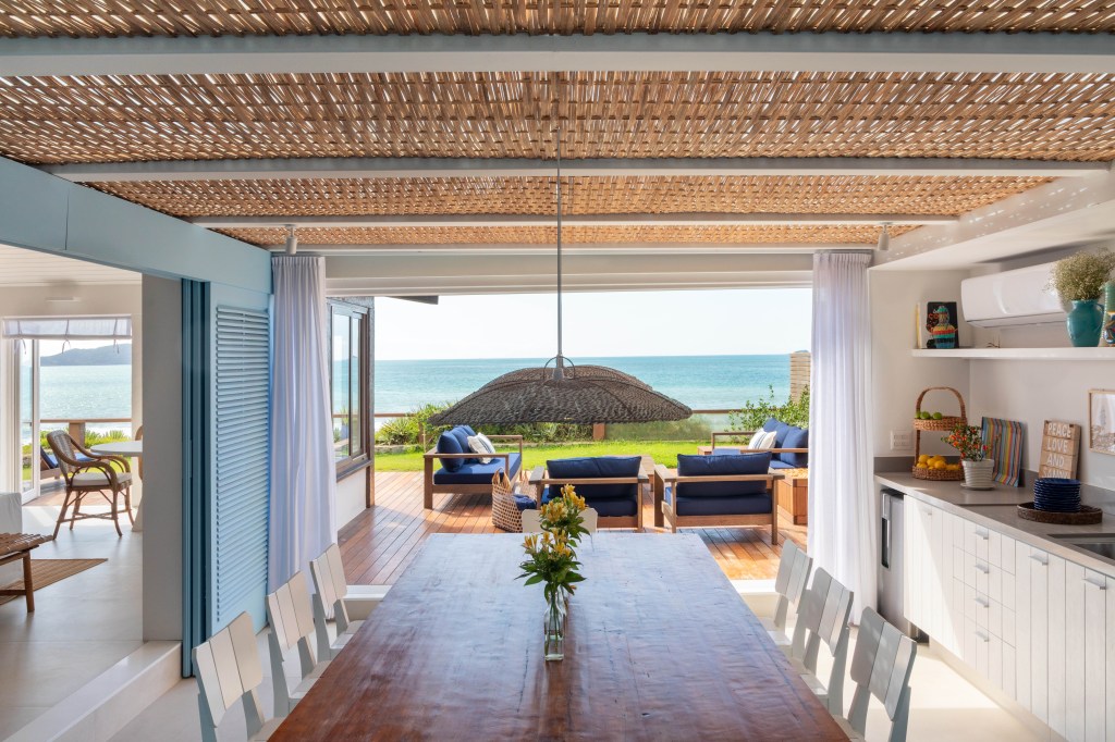 Venezianas azuis na varanda marcam esta casa de praia inspirada na Grécia. Projeto de Brise Arquitetura. Na foto, cozinha gourmet com teto de palha e vista para o mar.