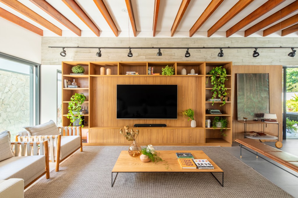 Pensada para veraneio, casa em meio ao verde vira o lar oficial da família. Projeto de Pietro Terlizzi. Na foto, sala de tv com estante de madeira e ripas no teto.