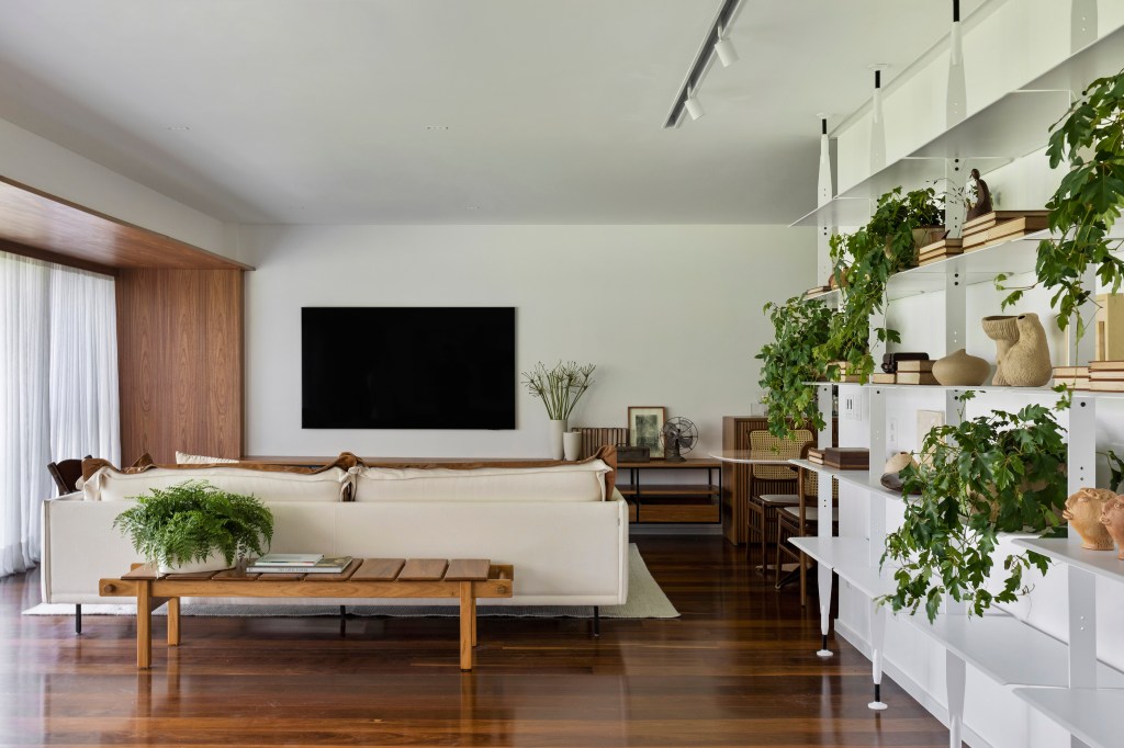 Com ampla varanda, apê de 200 m² tem décor inspirada no bairro do Leblon. Projeto de João Panaggio. Na foto, sala com tv, prateleiras com plantas e piso de madeira.