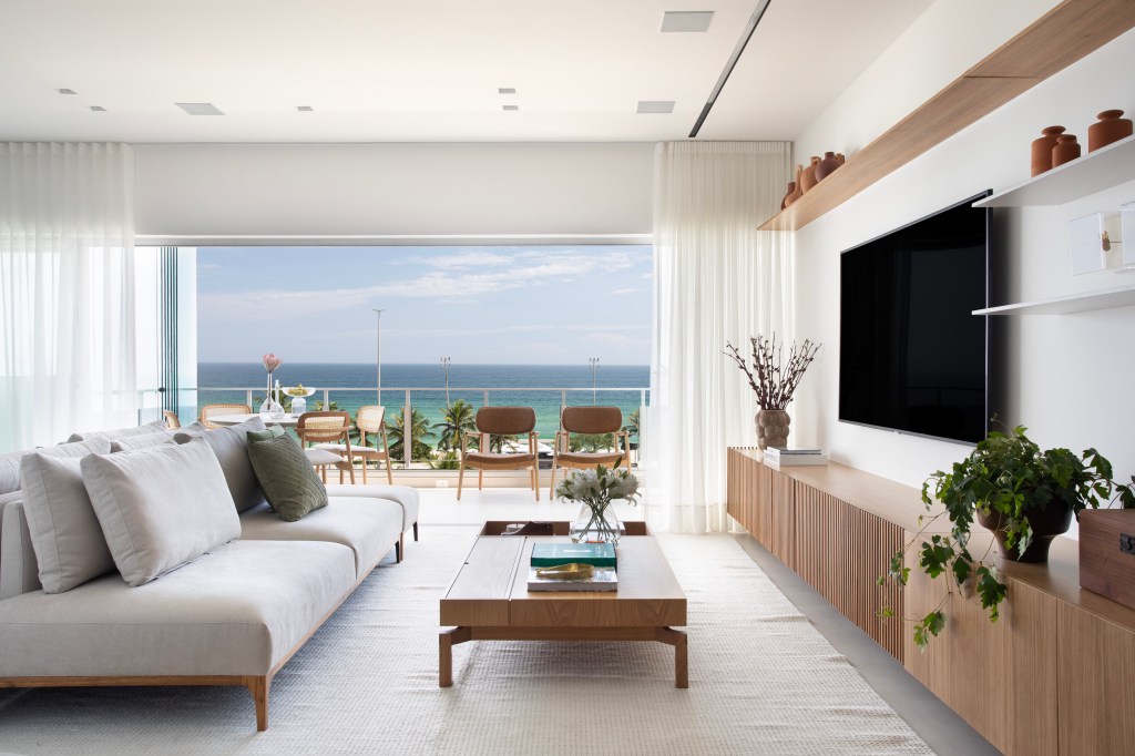 De frente para o mar, um apartamento ideal para curtir a aposentadoria. Projeto de Rafael Ramos. Na foto, sala de TV com varanda prateleiras e sofá.