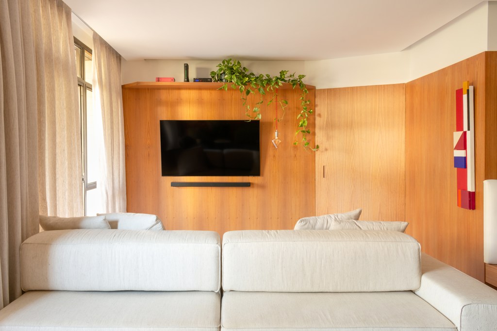 madeira-protagonista-apartamento-117-m2-gavea-mariana-monnerat-credito-lilia-mendel (9) A madeira é a protagonista deste apartamento de 117 m² na Gávea. Projeto de Mariana Monnerat, Na foto, sala com tv, painel de madeira e sofá ilha.