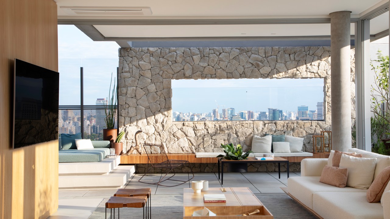 Skyline de São Paulo é emoldurado em apê assinado por Sabugosa Arquitetura. Na foto, Sala de estar integrada à varanda com muro de pedra e vista de SP.