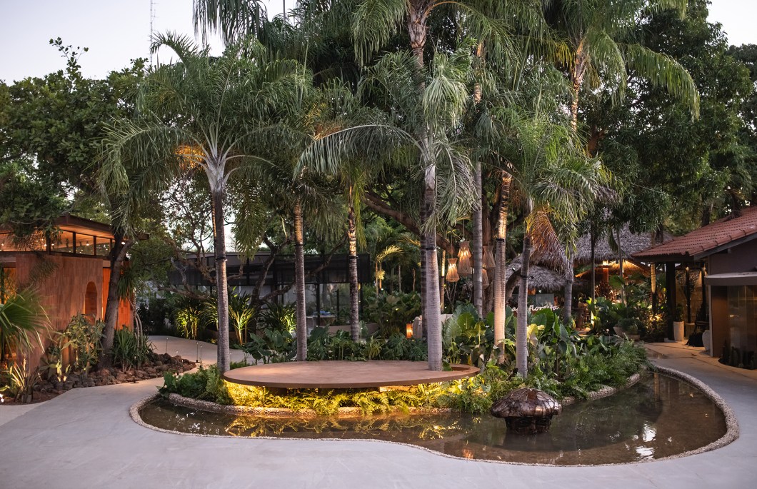 <b>Josélia Carvalho Paisagismo e Godoy Arquitetos - Praça Dois Rios.</b> As palmeiras majestosas que foram mantidas do jardim original da casa são guardiãs silenciosas, testemunhas do passado e vigias do presente. Ao redor delas, a lâmina d'água sinuosa, dividida em dois níveis, evoca os fluxos dos rios ancestrais do Piauí: o Poti e o Parnaíba. Os lounges oferecem refúgio do tumulto, convidando à contemplação tranquila da vegetação composta por plantas tropicais e espécies nativas. O mobiliário selecionado é uma celebração do conforto e da funcionalidade, incentivando os visitantes a se acomodarem e se entregarem ao prazer da companhia e da conversa descontraída. <b>Josélia Carvalho Paisagismo e Godoy Arquitetos - Praça Dois Rios.</b> As palmeiras majestosas que foram mantidas do jardim original da casa são guardiãs silenciosas, testemunhas do passado e vigias do presente. Ao redor delas, a lâmina d'água sinuosa, dividida em dois níveis, evoca os fluxos dos rios ancestrais do Piauí: o Poti e o Parnaíba. Os lounges oferecem refúgio do tumulto, convidando à contemplação tranquila da vegetação composta por plantas tropicais e espécies nativas. O mobiliário selecionado é uma celebração do conforto e da funcionalidade, incentivando os visitantes a se acomodarem e se entregarem ao prazer da companhia e da conversa descontraída.