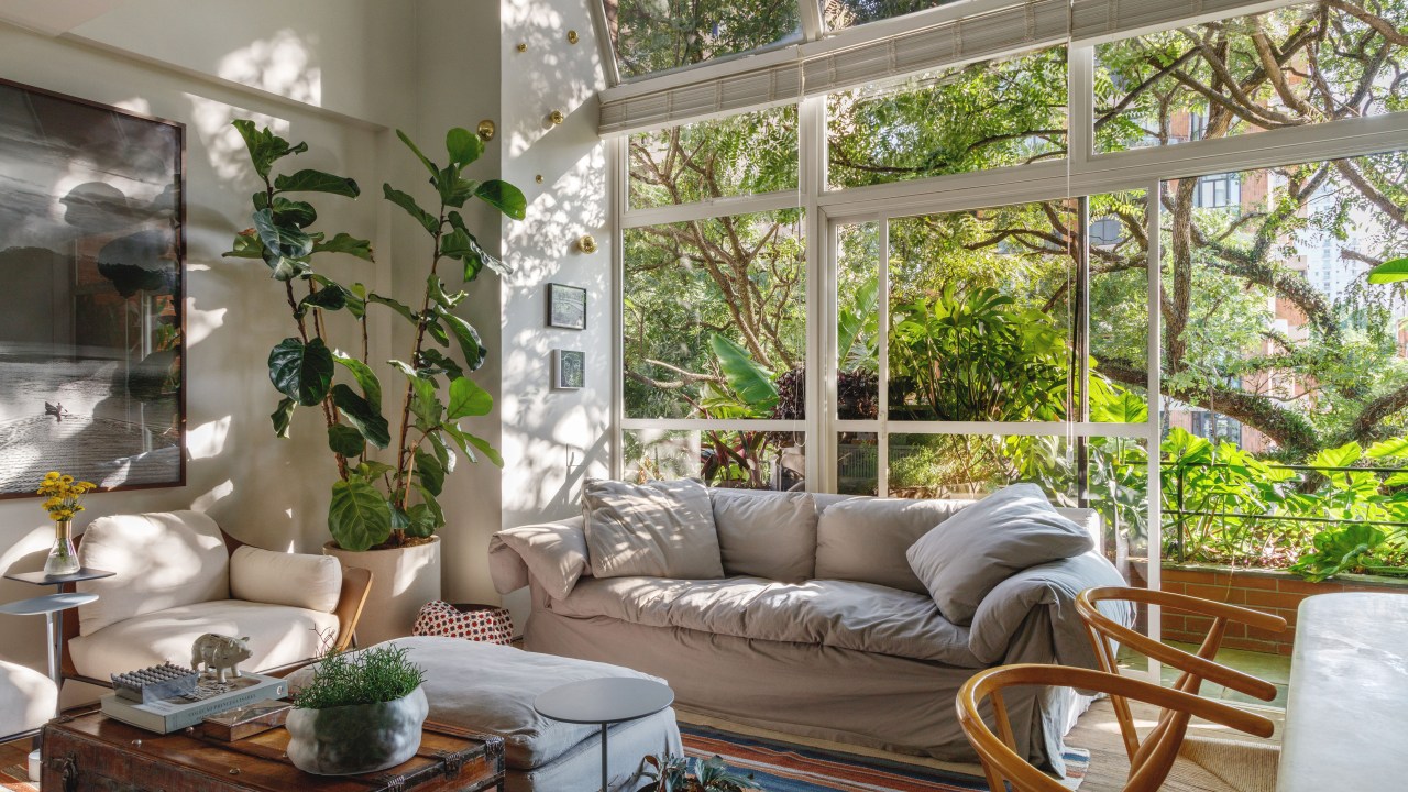 Aberturas de vidro piso-teto trazem árvores para dentro deste apê de 95 m². Projeto de DCC Arquitetura. Na foto, sala com parede de vidro, varanda com plantas e poltrona.