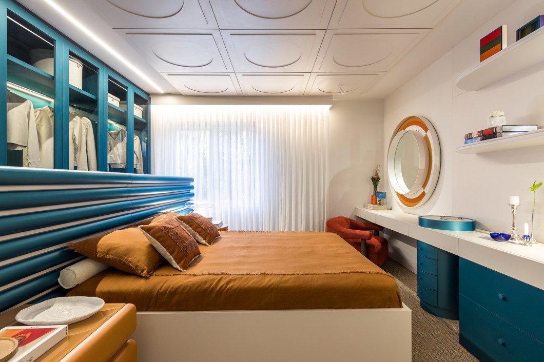 <b>Flávia Bonet - Quarto do Casal.</b> Um quarto que transporta os visitantes às memórias afetivas dos anos 1980, década que marca o nascimento do casal descolado e de alma jovem que inspira o ambiente. A marcenaria embutida, típica da arquitetura da época, garante o máximo aproveitamento do pequeno espaço. Elementos curvos e coloridos comportam os objetos trazidos das aventuras vivenciadas mundo afora pela dupla - que ama viagens e cores. Tons fortes e vibrantes, como laranja e azul, se somam aos acabamentos metalizados da laca de alto brilho para remeter ao cenário alegre e divertido que marca esta geração. A ambientação é arrematada por peças assinadas, itens colecionáveis e obras de artistas locais, todos inspirados em designers americanos que seguem o estilo “happy chic”. <b>Flávia Bonet - Quarto do Casal.</b> Um quarto que transporta os visitantes às memórias afetivas dos anos 1980, década que marca o nascimento do casal descolado e de alma jovem que inspira o ambiente. A marcenaria embutida, típica da arquitetura da época, garante o máximo aproveitamento do pequeno espaço. Elementos curvos e coloridos comportam os objetos trazidos das aventuras vivenciadas mundo afora pela dupla - que ama viagens e cores. Tons fortes e vibrantes, como laranja e azul, se somam aos acabamentos metalizados da laca de alto brilho para remeter ao cenário alegre e divertido que marca esta geração. A ambientação é arrematada por peças assinadas, itens colecionáveis e obras de artistas locais, todos inspirados em designers americanos que seguem o estilo “happy chic”.